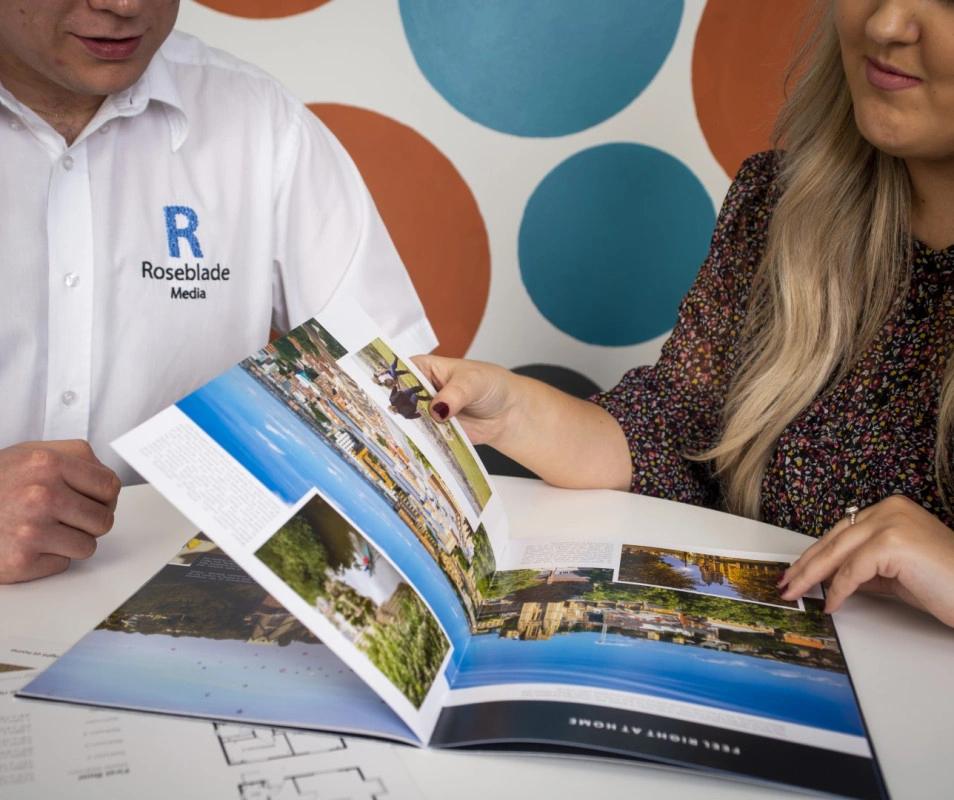 Two people are sitting at a table, looking at a brochure with landscape photographs, potentially discussing media or design work. (Captioned by AI)